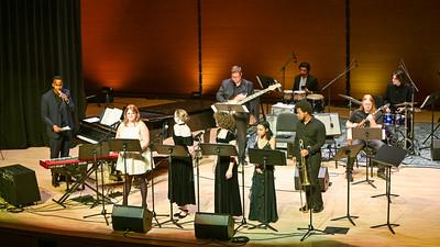 A jazz ensemble performs on stage with vocalists and instrumentalists, including a pianist, drummer, keyboardist, trombonist, and saxophonist, under warm stage lighting.