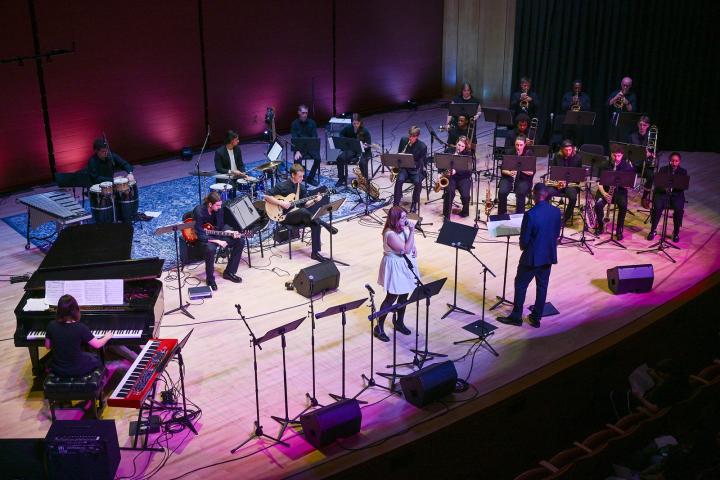 A large jazz band with multiple musicians performs on stage, featuring a singer and conductor in the front center, with various instruments including piano, drums, and brass.