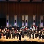 A large choir perform on stage with a conductor, standing in front of a wood-paneled backdrop with vertical decorations, under stage lighting.