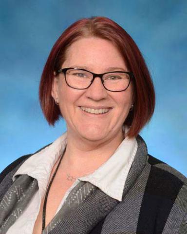A woman with short, straight, reddish hair and glasses smiles at the camera. She is wearing a white collared shirt and a patterned jacket, with a blue background behind her.