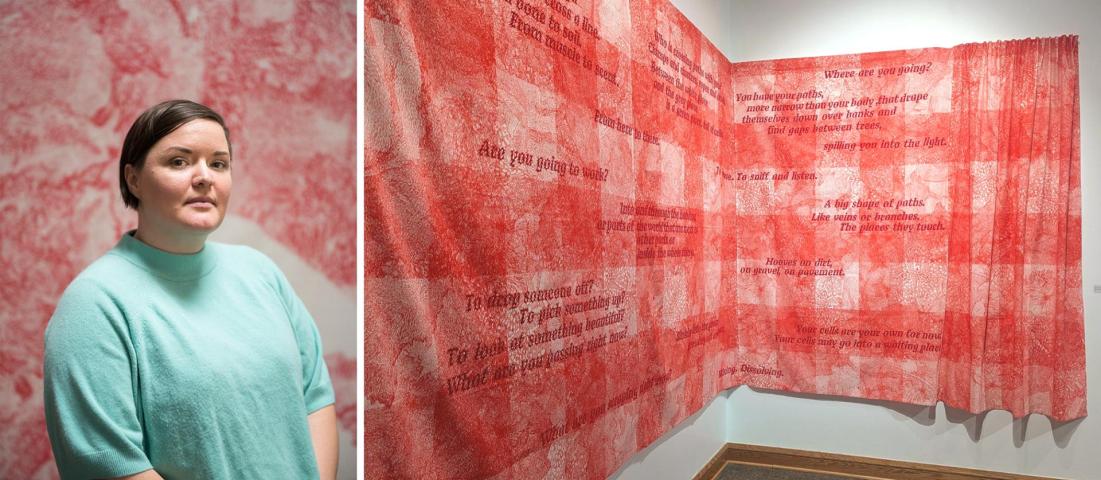 A person in a teal top stands beside a red and white textile artwork with text displayed on a wall in a gallery setting.
