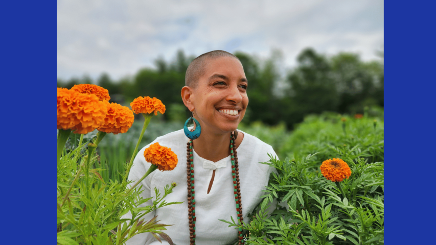 A Leah Penniman  wearing a white shirt and beaded necklace, smiles among orange marigold flowers in a green garden.
