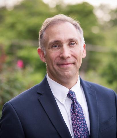 A man wearing a dark suit, white shirt, and patterned tie stands outdoors in front of greenery, smiling slightly at the camera.