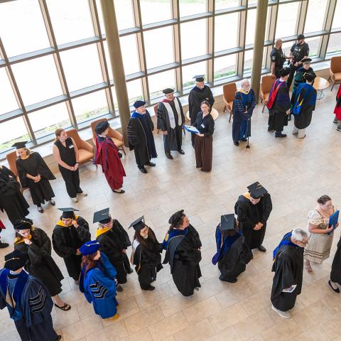 A group of faculty members in academic regalia stand in a sunlit hall, some talking and others waiting in a line.