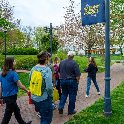 A group of people walking along a brick path on a college campus with a banner that reads “The National Public Honors College.”