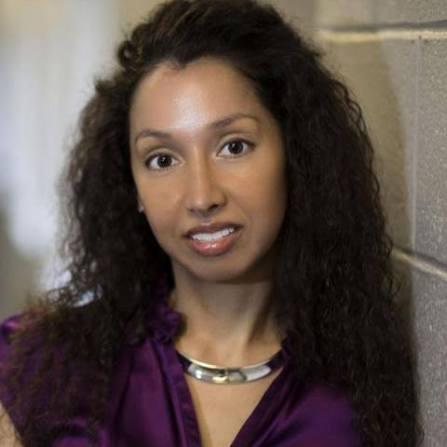 A woman with long, dark, curly hair wearing a purple top and a silver collar necklace.
