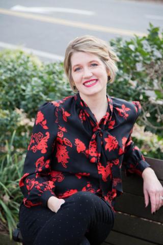 Neiles sits on a bench wearing a black shirt with red flowers, black pants, and black high heels.