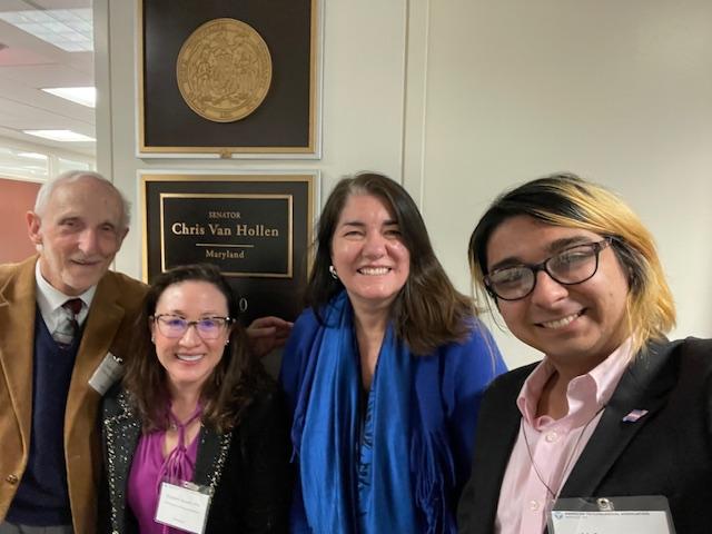 Four people are smiling and standing in front of a wall plaque for Senator Chris Van Hollen, Maryland. The group includes one person with glasses and another wearing a blue scarf. They appear to be in an office setting.