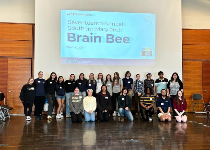 A group of 28 people stand and kneel in rows on a stage, smiling, in front of a large screen displaying text: "Seventeenth Annual Southern Maryland Brain Bee, March 1, 2025