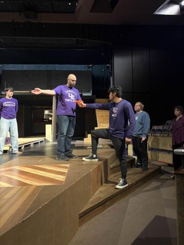 People on stage rehearsing. One person stands on a platform holding a script, while another person reaches out to them. Others watch nearby.