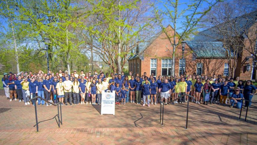 A large group of people standing outside in front of a brick building, gathered around a "Volunteer Check-In" signpost on a sunny day.