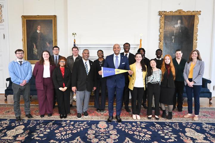 A group of people, dressed in business attire, stand together in a formal room with paintings on the walls. One person in the center holds a St. Mary's sign.