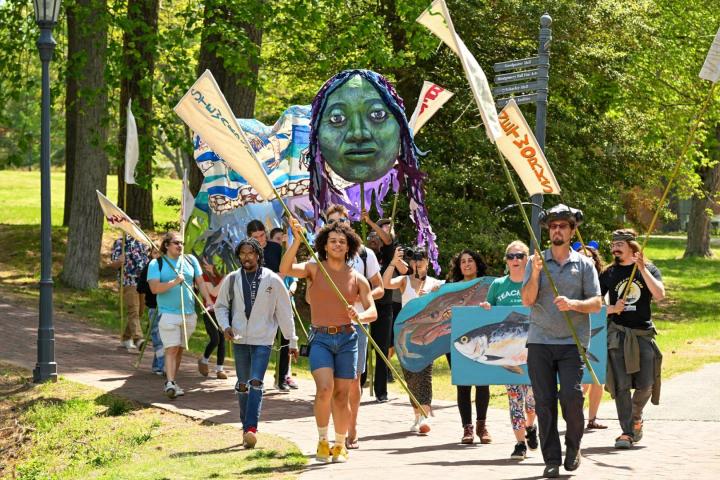 A group of people march outdoors holding banners, flags, a large puppet head, and a cardboard fish, with trees and greenery in the background.