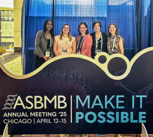 Five people stand behind an ASBMB Annual Meeting 2025 sign in Chicago, featuring the event theme "Make It Possible."