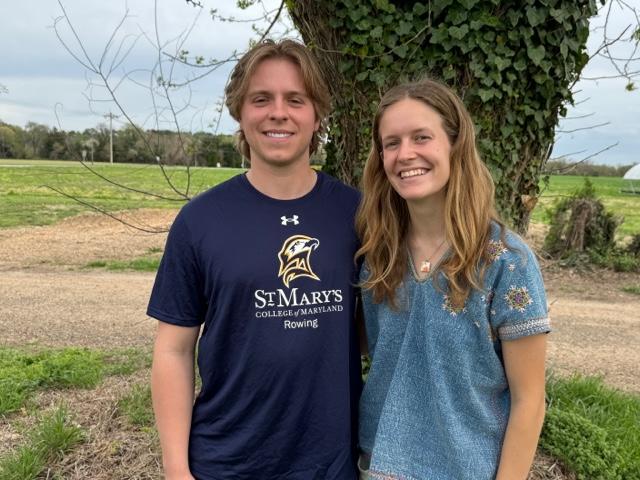 Two people standing outdoors, smiling at the camera. One wears a St. Mary's College rowing shirt. Green field and tree in the background.
