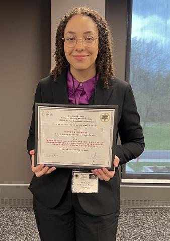 A person wearing glasses and a suit stands indoors holding a framed certificate and smiling at the camera.