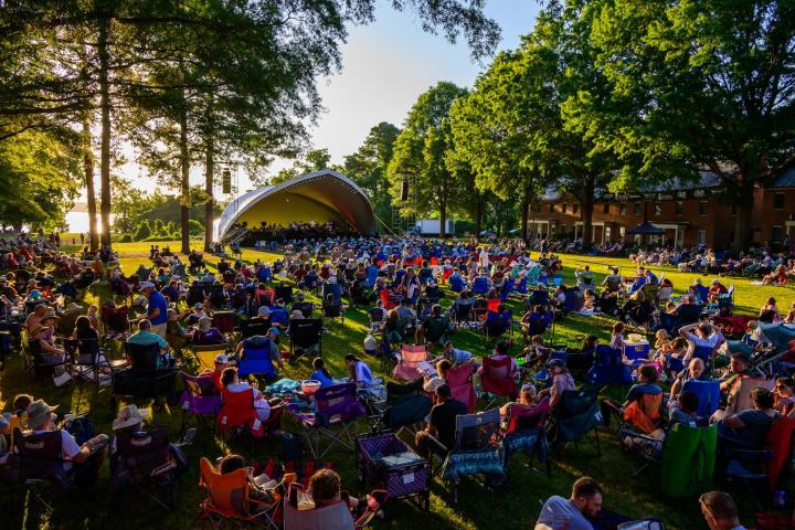 Large crowd sitting on lawn chairs and blankets at an outdoor concert in a park, with a stage set up under trees on a sunny day.