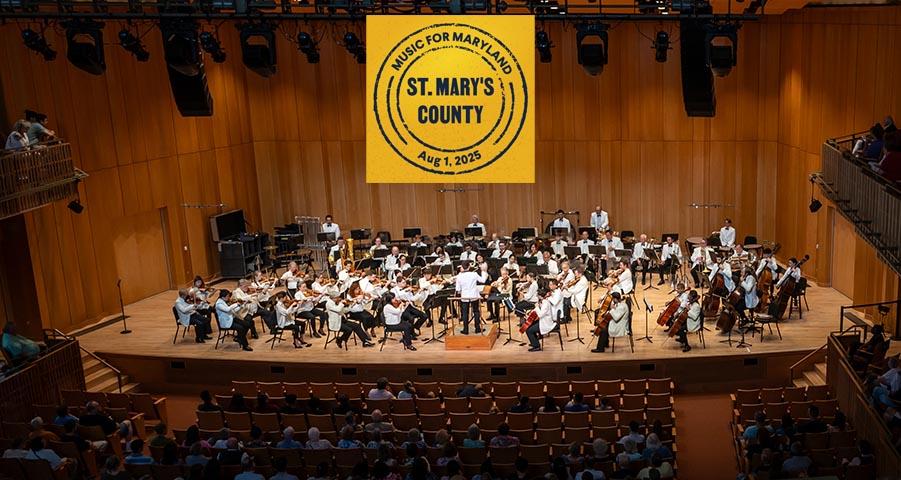 A symphony orchestra performs on stage in a wooden concert hall with a banner overhead reading "Music for Maryland, St. Mary's County, Aug. 1, 2025." Audience members are seated below.
