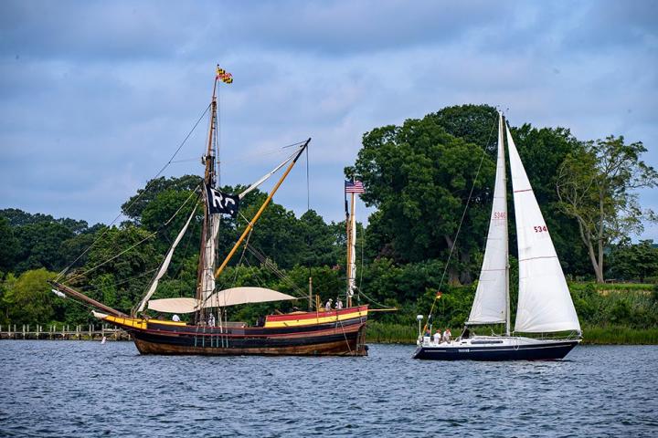 A wooden tall ship with a race committee flag and a modern white sailboat are side by side on a river, with trees and greenery in the background.