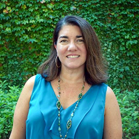 A woman with shoulder-length brown hair, wearing a blue sleeveless top and a beaded necklace, stands outdoors in front of a leafy green wall.