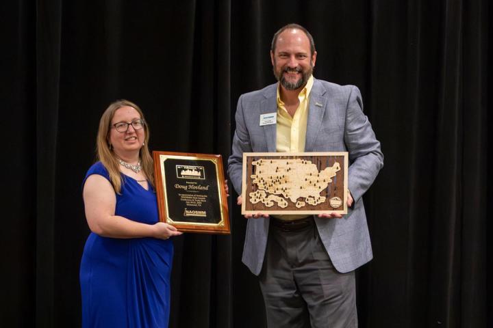 Two people stand in front of a black curtain including Doug Hovland; one holds a plaque, and the other holds a wooden map of the United States.