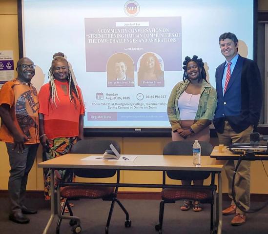 Four people stand in front of a projector screen displaying information about a community conversation event at Montgomery College. A table with chairs is in the foreground.