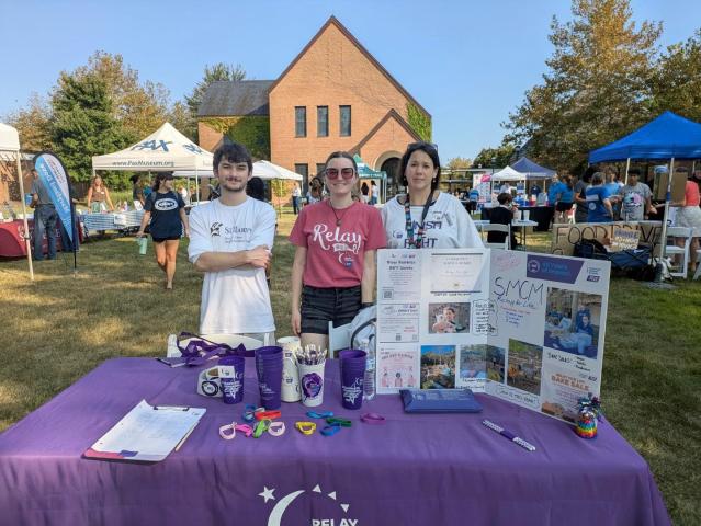 Three people stand behind a Relay for Life booth at an outdoor event, with informational materials and promotional items displayed on the table. Tents and people are visible in the background.