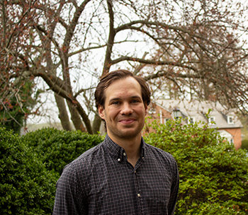 A man stands outdoors in front of green bushes and leafless trees, wearing a checked button-up shirt, smiling at the camera.