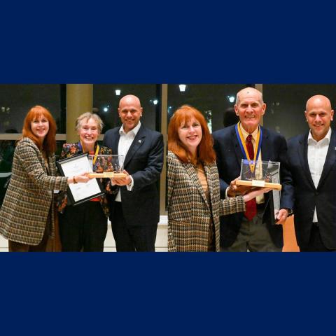 Six people stand indoors, with two women and one man holding awards and certificates, all dressed in formal or business attire, smiling at the camera.
