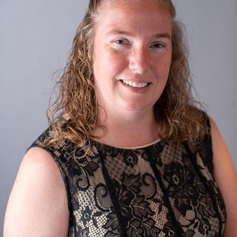 Woman with wavy, shoulder-length hair wearing a black lace dress, smiling, and posed against a plain gray background.