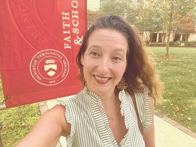 A woman smiles for a selfie in front of a Princeton Theological Seminary banner outdoors on a sunny day.