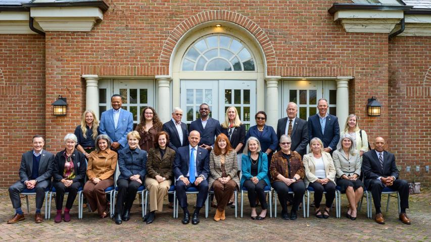 A group of 24 adults pose for a formal group photo in two rows outside a brick building with arched windows and columns.