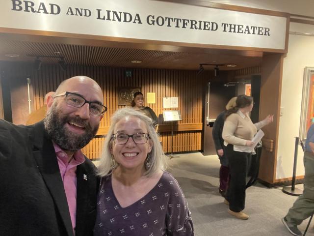 Two people smiling for a selfie inside a theater lobby with a sign reading “Brad and Linda Gottfried Theater” above them; several others are in the background.