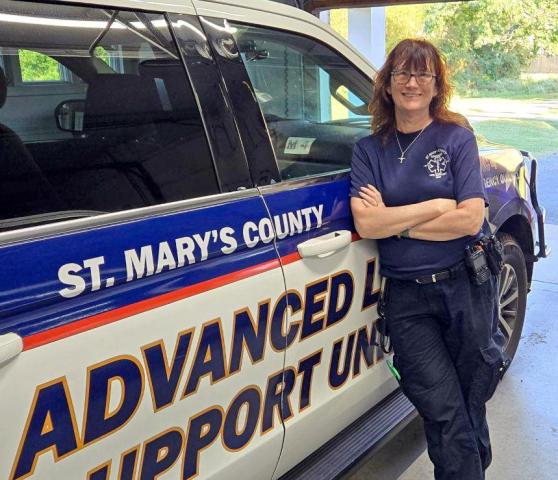 A person in a navy uniform stands smiling and leaning against a St. Mary's County Advanced Life Support Unit vehicle, parked inside a garage.