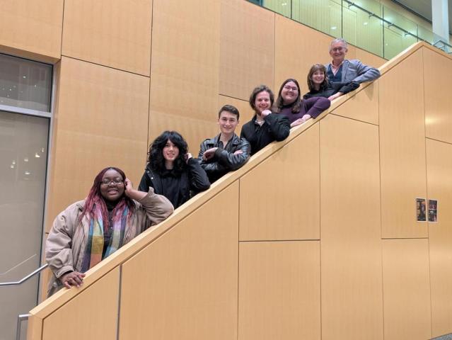 Seven people pose on a staircase inside a building with tan walls; six stand on the steps while one stands at the base, all facing the camera.
