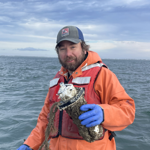 A man in an orange life jacket and blue gloves stands on a boat holding a barnacle-covered object over the water under a cloudy sky.
