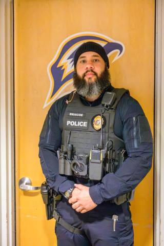 A police officer wearing a tactical vest stands in front of a wooden door with a sports team logo.