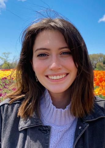 A woman with shoulder-length brown hair smiles outdoors in front of colorful flowers under a blue sky.