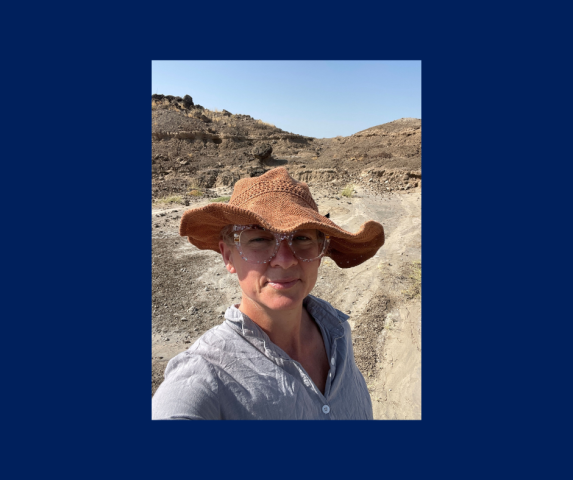 A person wearing a wide-brimmed hat, glasses, and a light shirt stands outdoors on a dusty path with rocky terrain in the background under a clear sky.