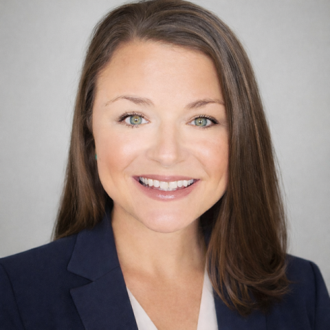 A woman with straight brown hair, wearing a navy blazer and light blouse, smiles at the camera against a plain light gray background.