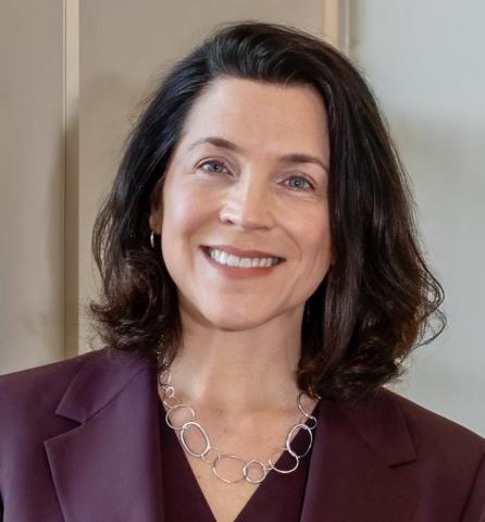 A woman with shoulder-length dark hair, wearing a maroon blazer and a silver chain necklace, smiles at the camera against a neutral background.