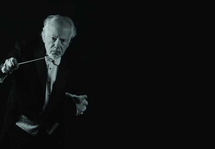 An older man in formal attire holds a conductor’s baton and poses against a dark background, appearing to conduct music.