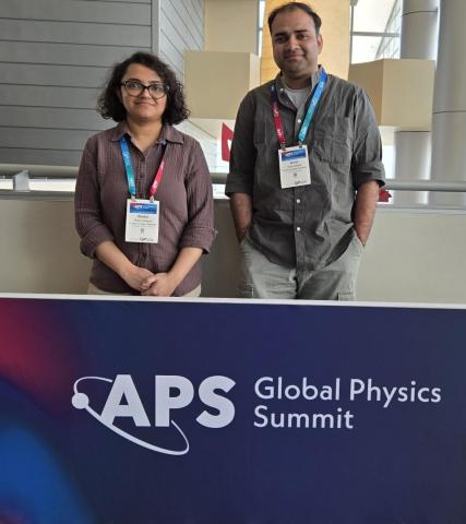 Drs. Shalini Ganguly (left) and Anish Agashe (right) stand behind a banner that reads "APS Global Physics Summit," each wearing conference badges.