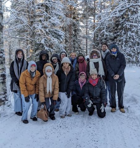 A group of fourteen people dressed in winter clothing pose for a photo on a snowy path surrounded by snow-covered trees.