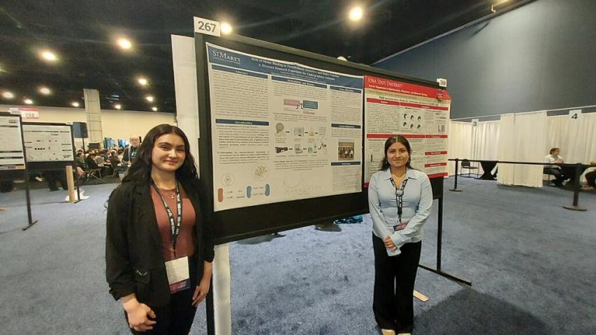 Two women stand in front of a scientific research poster at a conference, both smiling at the camera. The room is filled with other posters and attendees in the background.