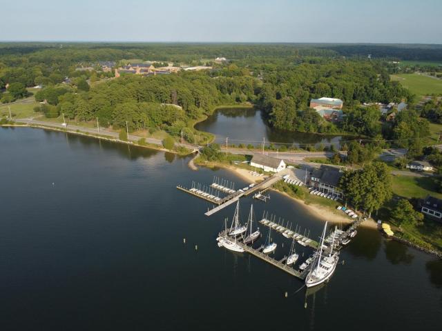 Aerial view of a marina with sailboats docked at piers, surrounded by shoreline buildings, trees, and a pond, with a forested area and fields in the background.