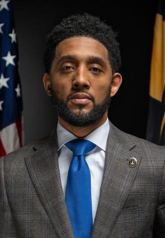 A man in a plaid suit jacket and blue tie stands in front of American and state flags, facing the camera with a neutral expression.