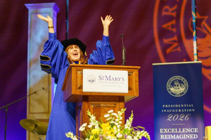 A person in academic regalia stands at a podium with arms raised, speaking at a St. Mary’s College of Maryland event, with a “Presidential Inauguration 2026” sign nearby.