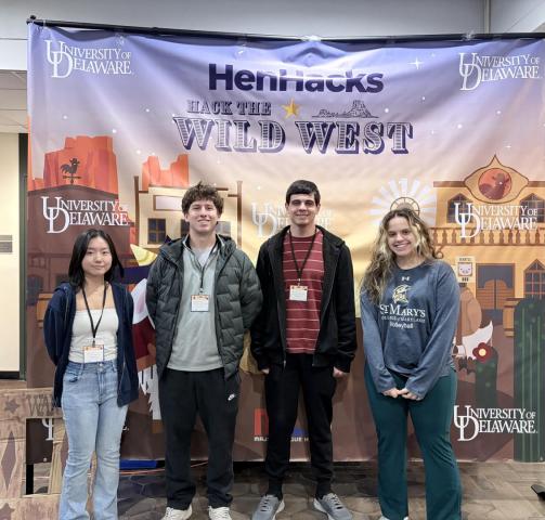 Four students stand in front of a "Hack the Wild West" HenHacks banner at the University of Delaware.