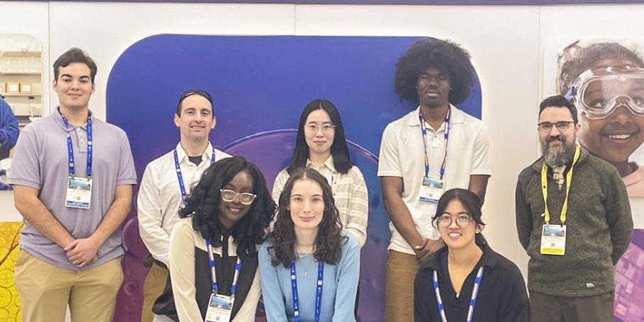 A group of eight people pose indoors in front of a colorful backdrop at what appears to be a conference or event, all wearing name badges.